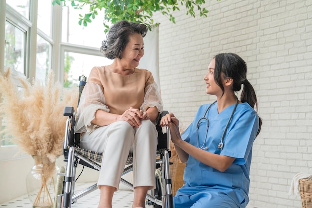 Asian young caregiver caring for her elderly patient at senior daycare Handicap patient in a wheelchair at the hospital talking to a friendly nurse and looking cheerful nurse wheeling Senior patient