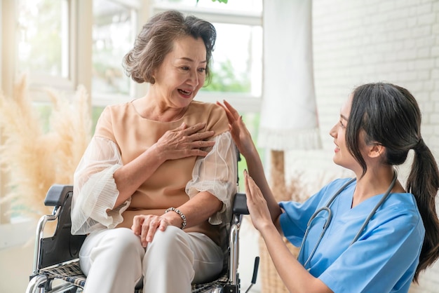 Asian young caregiver caring for her elderly patient at senior daycare Handicap patient in a wheelchair at the hospital talking to a friendly nurse and looking cheerful nurse wheeling Senior patient