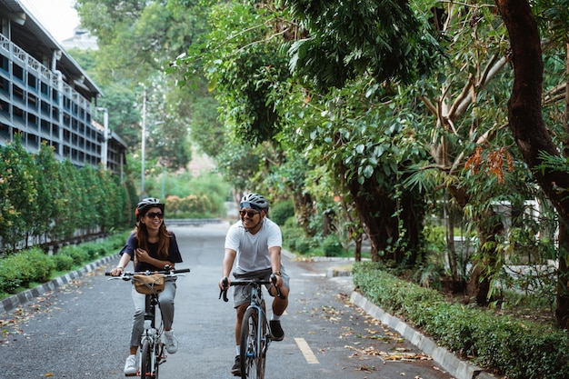 ヘルメットを身に着けているアジアの若いカップルは一緒に自転車に乗ることを楽しむ