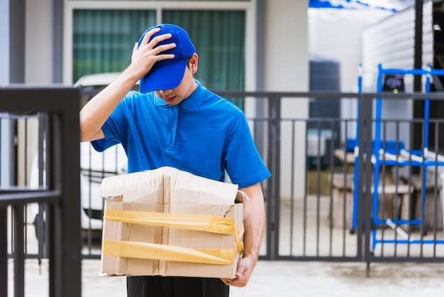 Asian young delivery man in blue uniform he emotional falling courier hold damaged cardboard box is broken at door front home, Accident bad transport shipment or poor quality delivery service concept