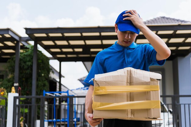 Asian young delivery man in blue uniform he emotional falling courier hold damaged cardboard box is broken at door front home, Accident bad transport shipment or poor quality delivery service concept