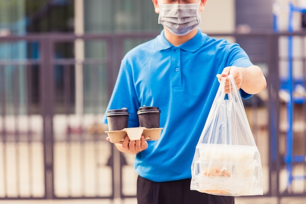 Asian young delivery man in blue uniform wearing face mask making grocery service giving rice food boxes plastic bags and coffee at front house under pandemic coronavirus, Back to new normal concept