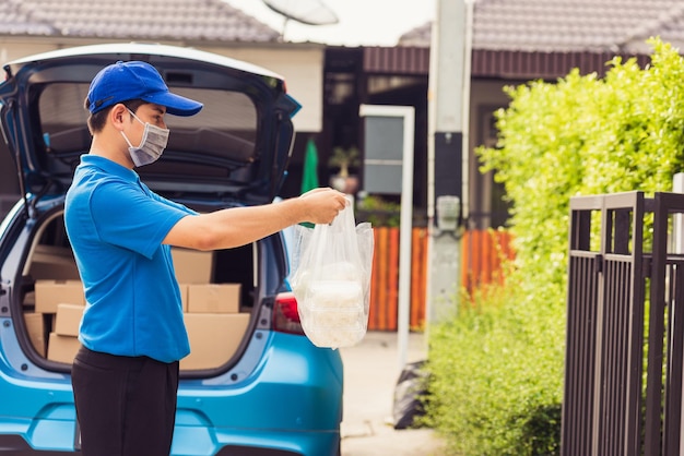 Asian young delivery man in blue uniform wearing face mask making grocery service giving rice food boxes plastic bags and coffee at front house under pandemic coronavirus, Back to new normal concept