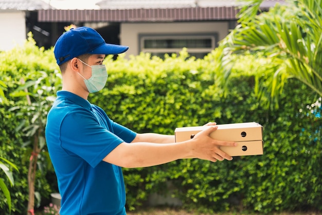 Asian young delivery man courier sending and holding fast food pizza boxed in uniform he protective face mask service customer at home door, under curfew quarantine pandemic coronavirus COVID-19