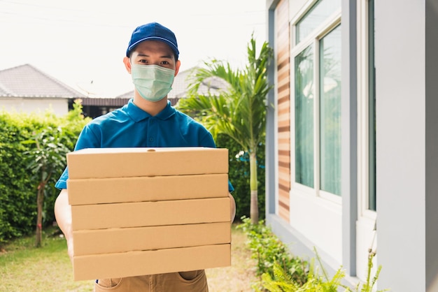 Asian young delivery man courier sending and holding fast food pizza boxed in uniform he protective face mask service customer at home door, under curfew quarantine pandemic coronavirus COVID-19