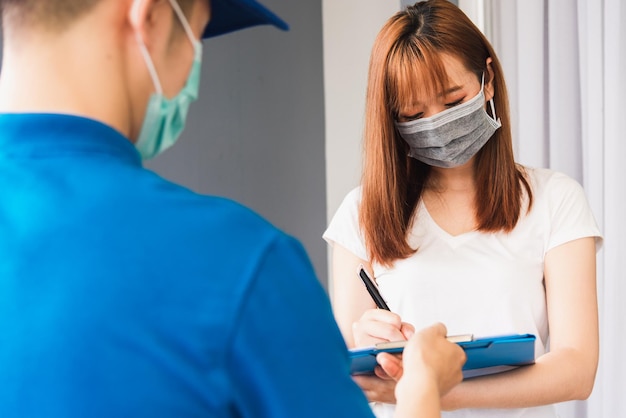 Asian young delivery man courier shopping online with package post box in uniform protective face mask coronavirus he making service woman customer using pen signature on paper book at home, COVID-19