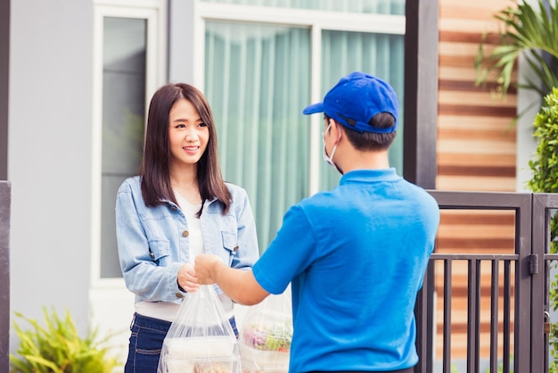 Asian young delivery man wear face mask he making grocery service giving rice food boxes plastic bags to woman customer receiving door at house after pandemic coronavirus, Back to new normal concept