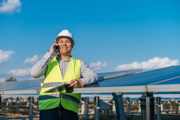 백그라운드에서 태양열 발전소에서 일하는 동안 손에 체크 보드를 들고 스마트폰으로 이야기하는 흰색 헬멧을 쓴 아시아 젊은 엔지니어