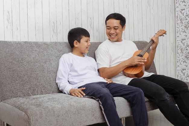 Asian young father in white shirt is playing ukulele while smiling to his son in white sweater spen