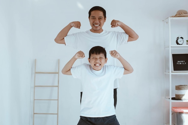 Asian young father in white t shirt and his son in white t shirt are smiling while pose showing musc