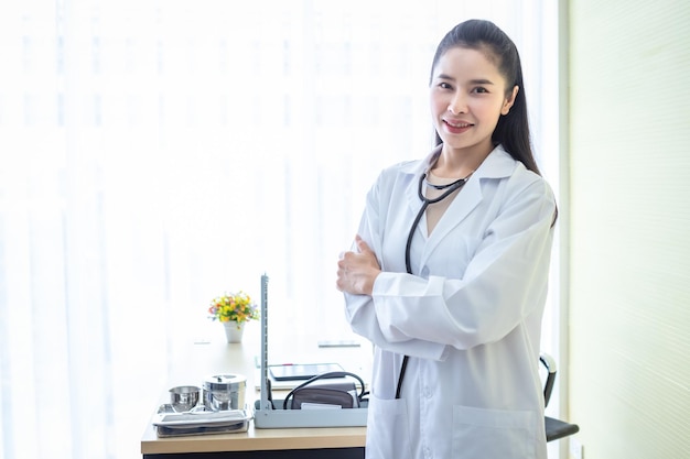 Asian young female doctor fold her arms with stethoscope with positive emotions in hospital background