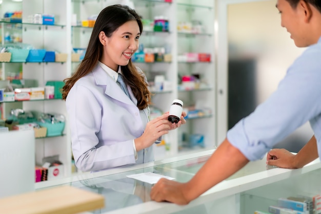 Азиатский молодой женский аптекарь с симпатичной дружелюбной улыбкой и объясняя медицину к ее клиенту в аптеке аптеки.