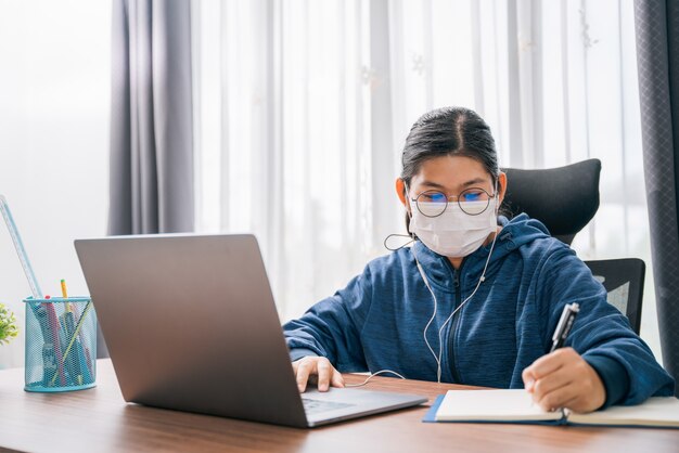 Asian young girl wear a mask student with glasses headphones\
girl study happy writing note on a book looking video conference\
laptop computer online internet learning distance education at\
home