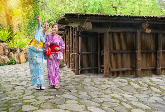 Giovani amiche asiatiche che indossano abiti kimono insieme puntando verso l'alto con la vista della natura e vicino alla vecchia casa di legno del giappone fuori durante le vacanze di viaggio al giorno del sole con copyspace.