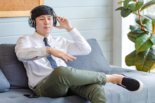 Азиатский молодой красивый профессиональный успешный бизнесмен-мужчина в формальной деловой рубашке и галстуке сидит на уютном диване и слушает потоковую музыку онлайн, просматривая интернет со смартфона.