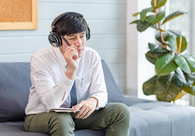 Asian young handsome professional successful male businessman employee in formal business shirt and necktie sitting on cozy sofa listening to streaming music online browsing internet with smartphone.