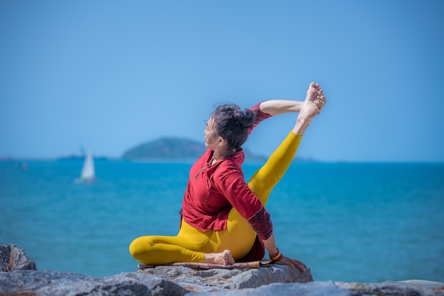 Photo asian young healthy woman posing practice bird of paradise yoga on stone beach with blue cloud sky