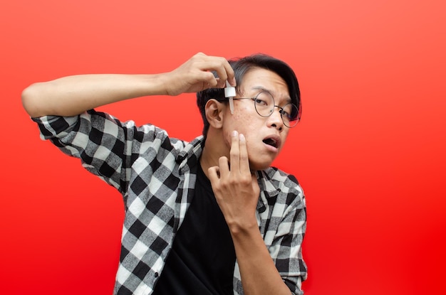 asian young man applying beauty acne serum and smiling at camera isolated over red background