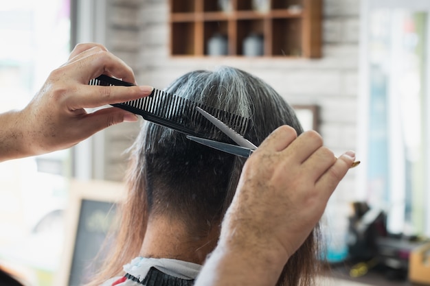 Asian young man being cut from long hair to short hair with scissor by professional barber in barbershop.