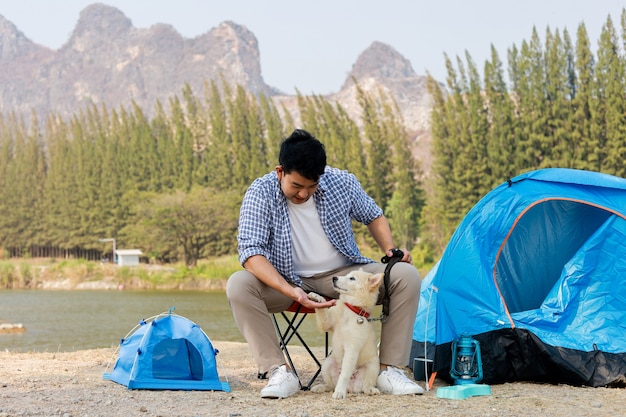 Photo asian young man in blue shirt with cute puppy dog camping on the lake hill mountain view happy and enjoy life