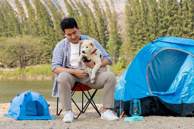 Il giovane asiatico in camicia blu con il cucciolo di cane sveglio che si accampa sul mountain view della collina del lago felice e gode della vita