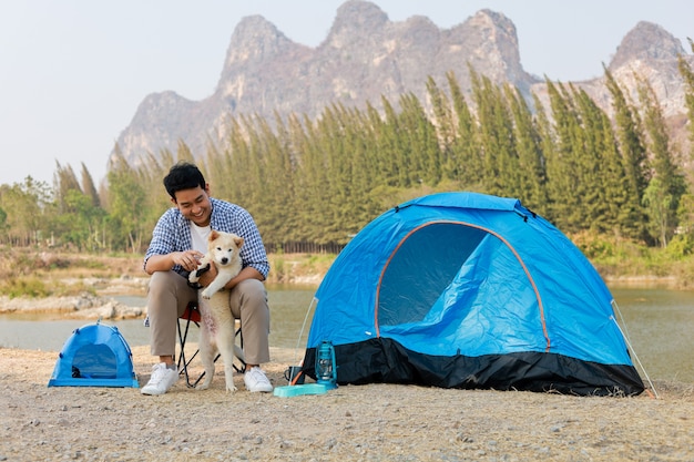 Asian young man in blue shirt with cute puppy dog camping on the lake hill mountain view happy and enjoy life