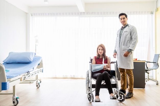 Asian Young man doctor of checking splint the arm of female patient hand due to with her arm broken for better healing with a smile sit in a wheelchair In the room hospital