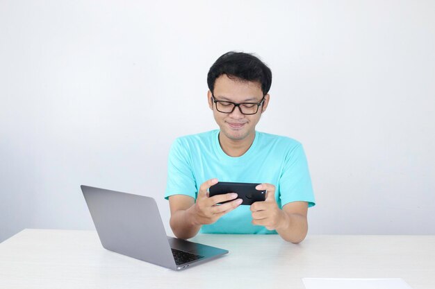 仕事でゲームをするときにスマートフォンで楽しむアジアの若い男青いシャツを着たインドネシア人の男