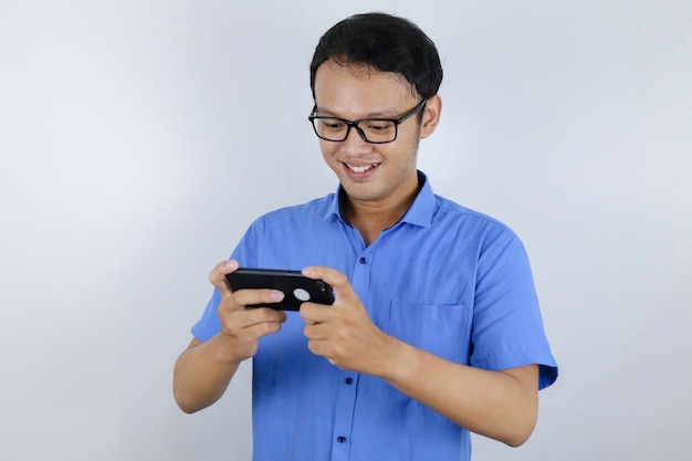 青いシャツを着たインドネシア人男性がゲームをプレイするとき、アジアの若い男性はスマートフォンで楽しんで笑顔を見せます