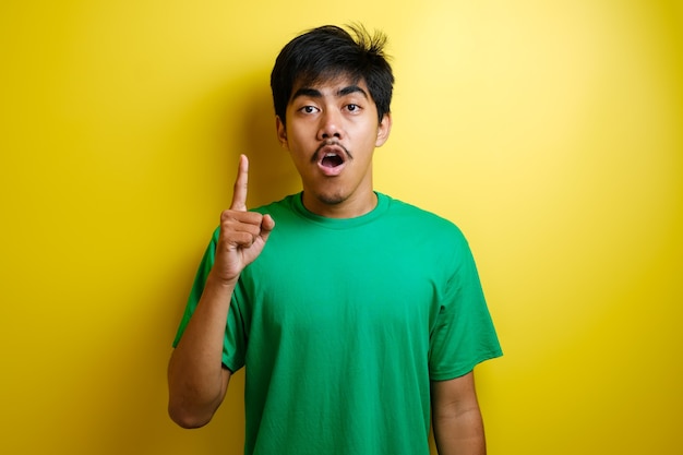 녹색 티셔츠를 입은 아시아 젊은이는 행복한 생각을 하고 위를 올려다보며 좋은 생각을 하고 있습니다. 복사 공간이 있는 노란색 배경의 반신 초상화