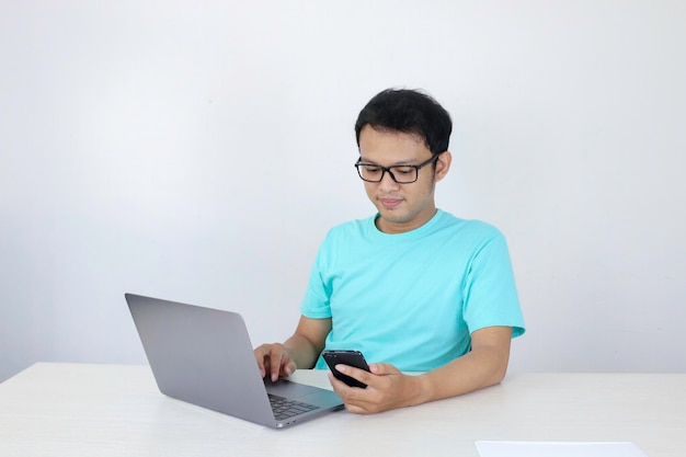 Asian young man happy and smile when he see on smart phone with laptop beside it Indonesian man wearing blue shirt