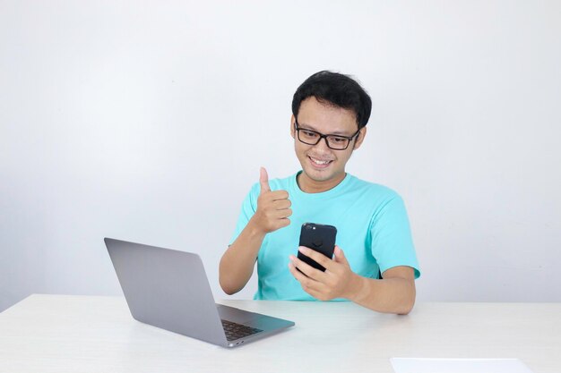 Asian young man happy and smile when he see on smart phone with laptop beside it Indonesian man wearing blue shirt