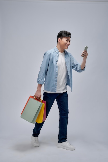 Asian young man holding shopping bags and using cellphone full length portrait isolated