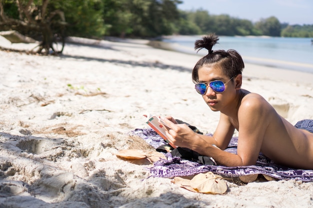 Giovane asiatico prendere il sole sulla spiaggia e leggere un libro