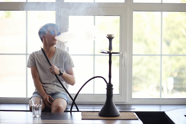 Asian young man in sunglasses smoking a hookah at the window