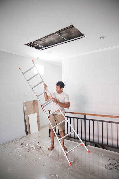 Asian young man using a ladder to repair the broken ceiling at\
home