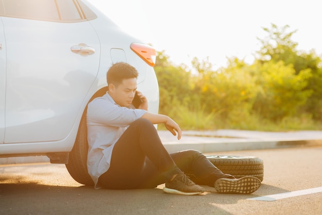 Asian young man using smartphone called a car repair shop or someone close to help after trying failed to fix a problem on the road Car broke down or changing flat tire on the road concept