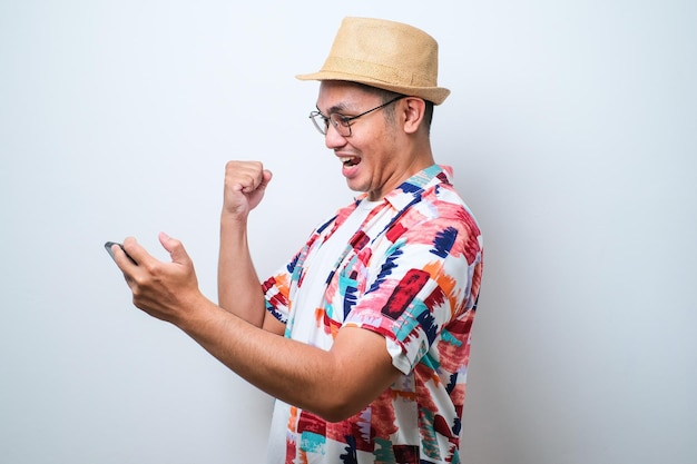 Asian young man wearing casual beach shirt holding smartphone doing winning gesture holding mobile phone