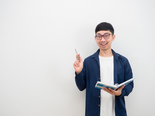 陽気な眼鏡をかけているアジアの若い男は、アイデア笑顔の顔のポイントペンを上げて本を持っています