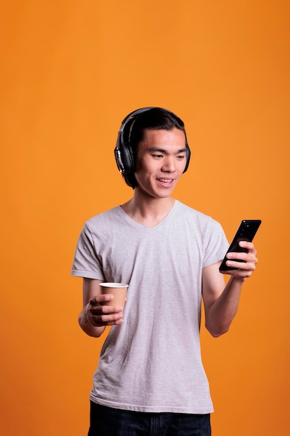 Asian young man in wireless headphones using social media on mobile phone, drinking take away coffee. Smiling teenager, listening to music on smartphone, holding tea paper cup
