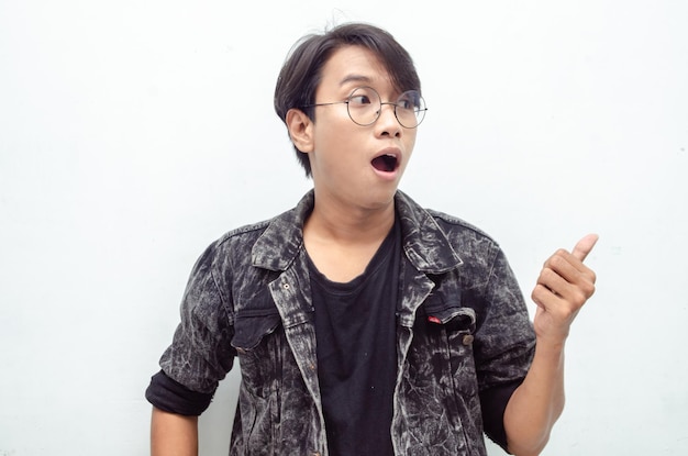 asian young man with glasses shocked happily pointing finger to copy space offering something goods