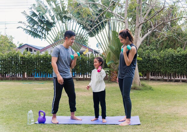 Asian young mother, father and child daughter doing exercising together with dumbbells is fun outdoors in nature a field garden park. Happy family kid sport and exercises for healthy lifestyle