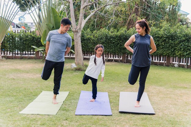 Asian young mother, father and child daughter practicing exercising stretching together before yoga workout outdoor morning in nature a field garden park. Happy family kid sport healthy lifestyle