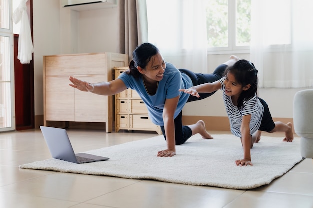 Asian young mother and her daughter doing stretching fitness exercise yoga together at home. Parent teaching child work out to be strong and maintain physical health and wellbeing in daily routine.