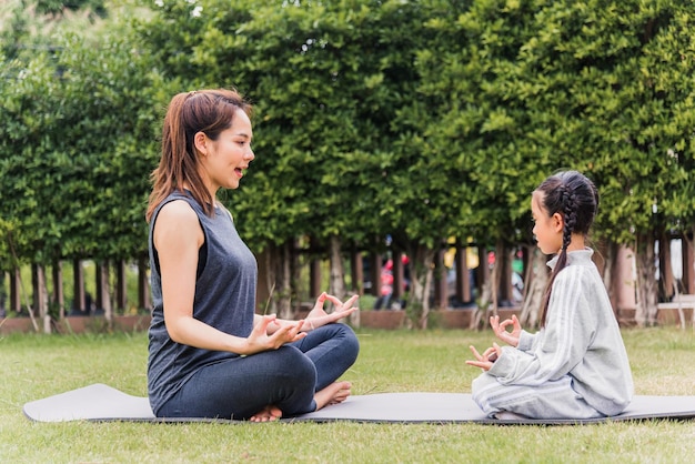 アジアの若い母親は、娘と一緒に屋外でヨガの練習をし、自然の中の緑の芝生の上で一緒に瞑想のポーズをとり、フィールドガーデンパーク、家族のスポーツ、健康的なライフスタイルのための練習をしています