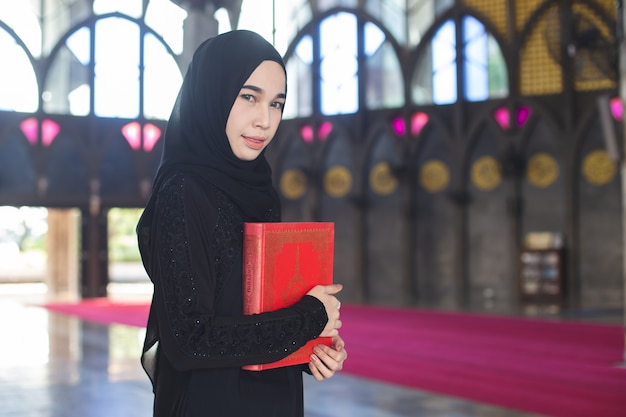 Giovane donna musulmana asiatica che tiene corano rosso, in moschea.