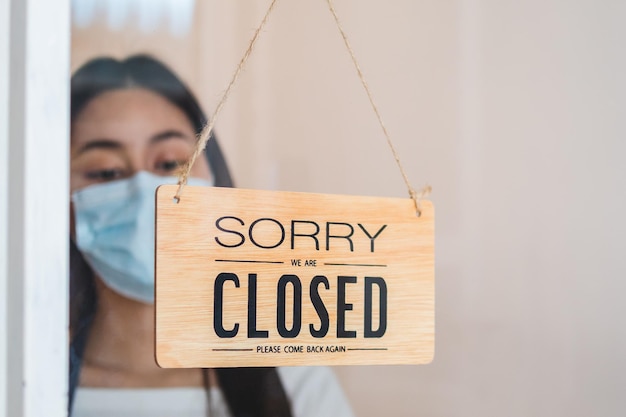 Foto giovane proprietario asiatico al dettaglio, donna della caffetteria che gira il cartello per chiudere con l'uso della maschera facciale, protezione dalla pandemia di coronavirus. chiudere negozio, ristorante o pub a causa del blocco, quarantena di covid.