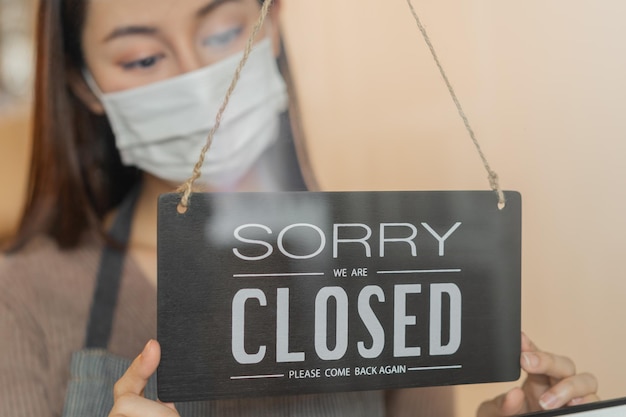 Asian young owner retail,coffee shop woman turning sign board to closed with wearing face mask,protection to pandemic of coronavirus.Close store,restaurant or pub due to lockdown, quarantine of covid.