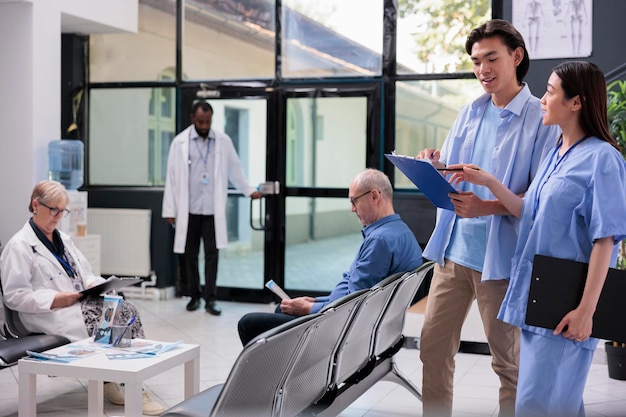 Asian young patient holding clipboard filling medical report\
before start consultation with medic during checkup visit in\
hospital waiting area. diverse people standing in reception,\
medicine service