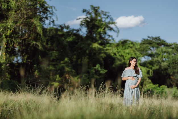 Asian young pregnant woman in blue dress relaxing and enjoying life in grass nature at summer day.